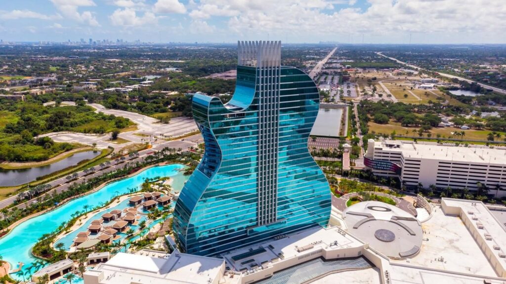 Aerial view of a guitar-shaped building with a vast pool complex, juxtaposed against a suburban backdrop.