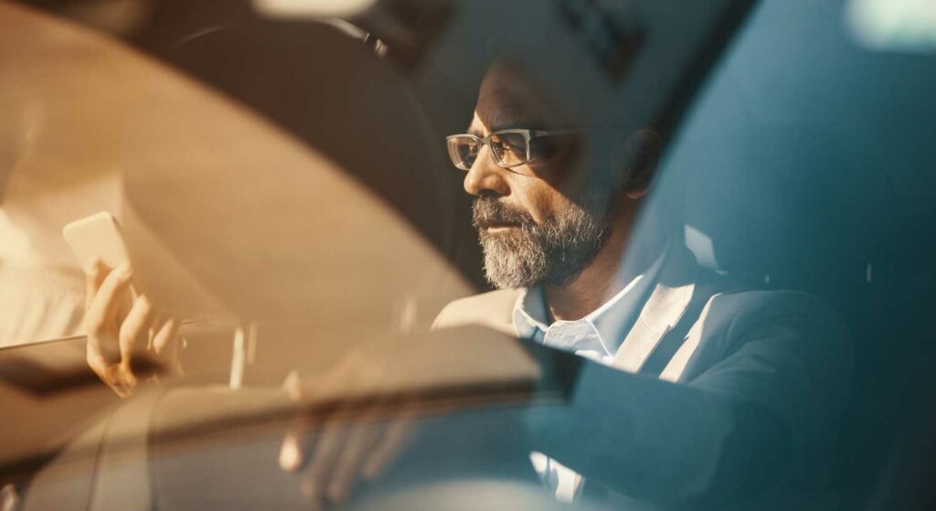 Mature businessman with glasses in the backseat of a car, looking contemplative while holding a smartphone.