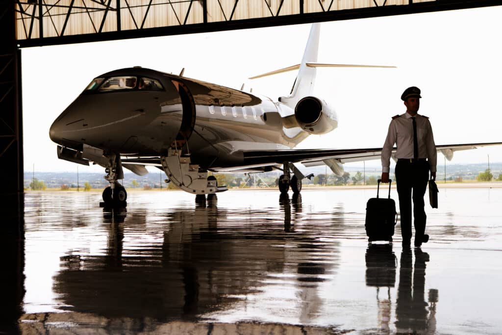 Private Jet, Older Male Pilot, wearing a pilot suit, white shirt, dragging a suitcase, walking towards the private jet, inside a private jet hanger.