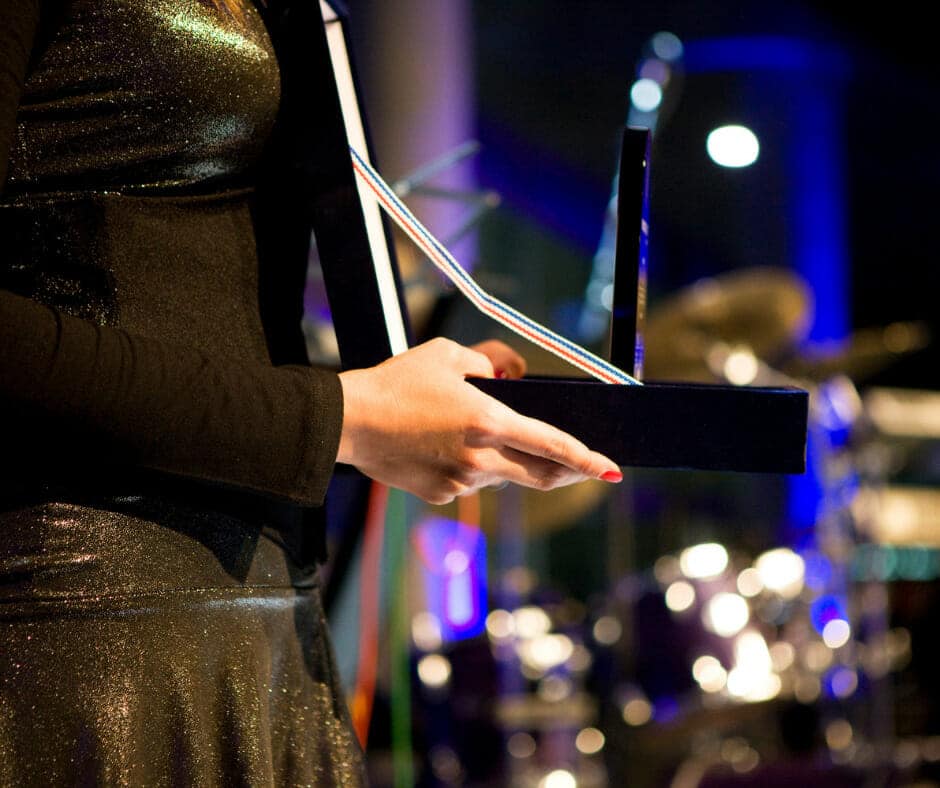 Close-up of a person's hands holding an award at an event, with a stage and musical instruments in the background.