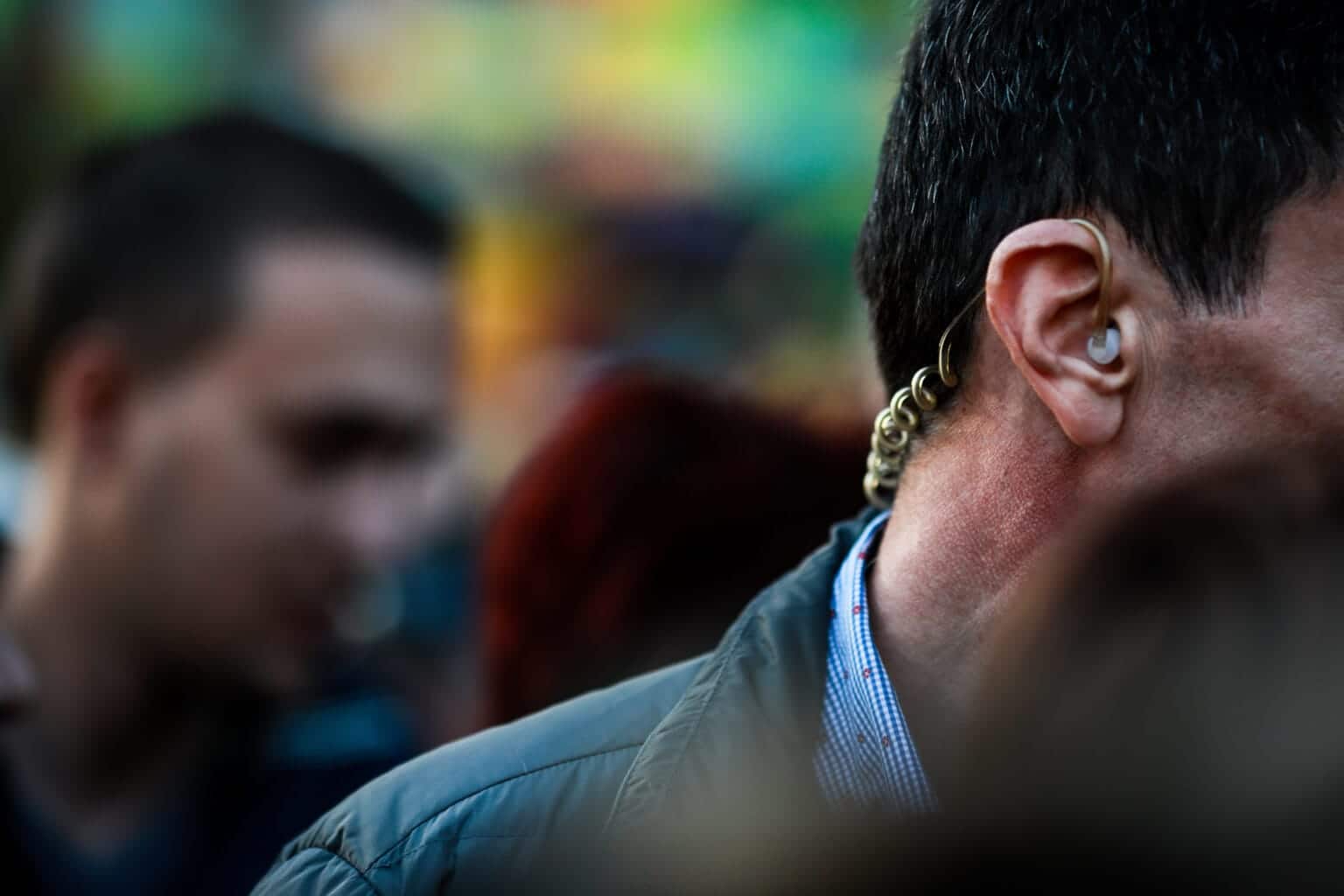 Close-up of a man's ear wearing a hearing aid with blurred faces in the background.