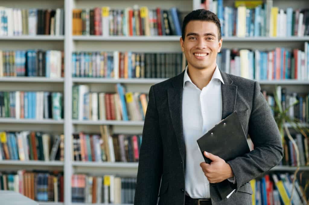 Confident young man holding a clipboard in a library with bookshelves in the background.