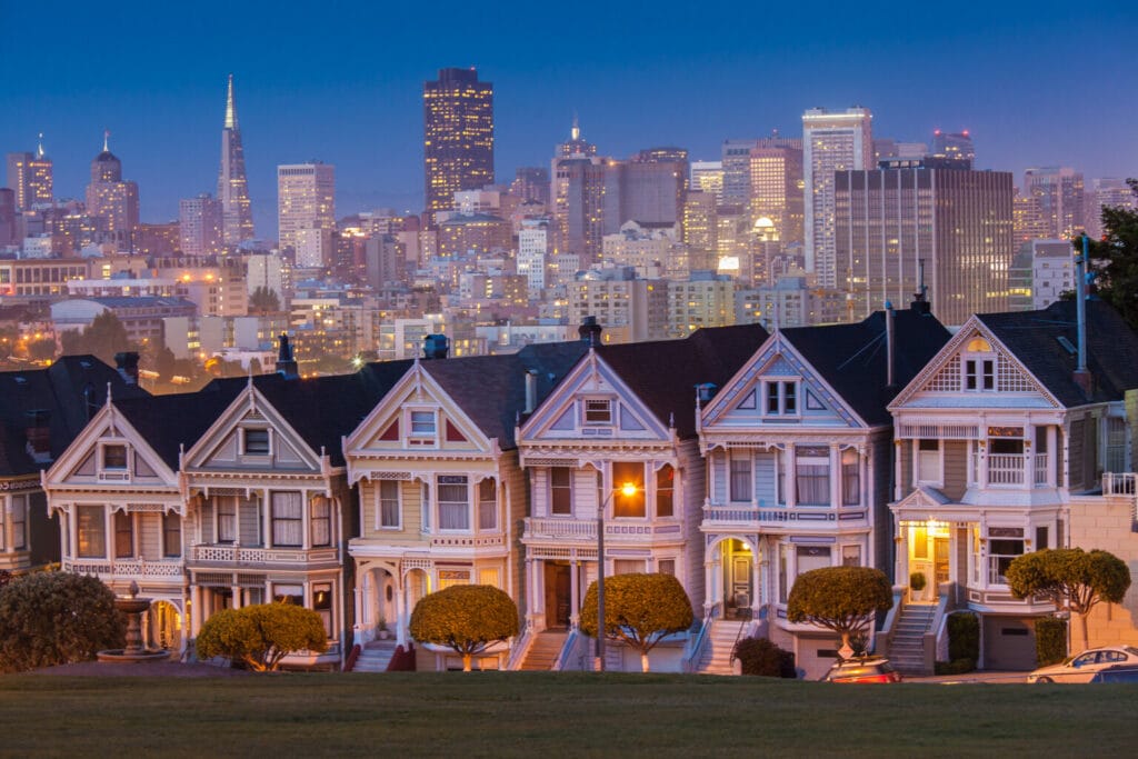 Historic Victorian houses in the foreground with a bustling city skyline behind at twilight.