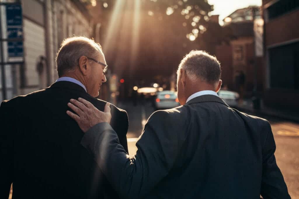 Two men in business suits walking outdoors, one with his hand on the other's back, in a gesture of guidance or support.