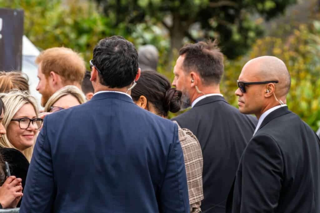 Team of security agents with earpieces standing among a crowd of people surrounding Prince Harry.