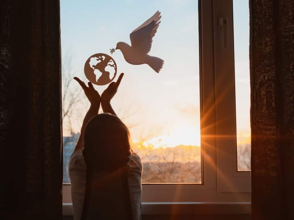 Silhouette of a person holding a cut-out of the Earth against a window, with a dove in flight and a sunset in the background