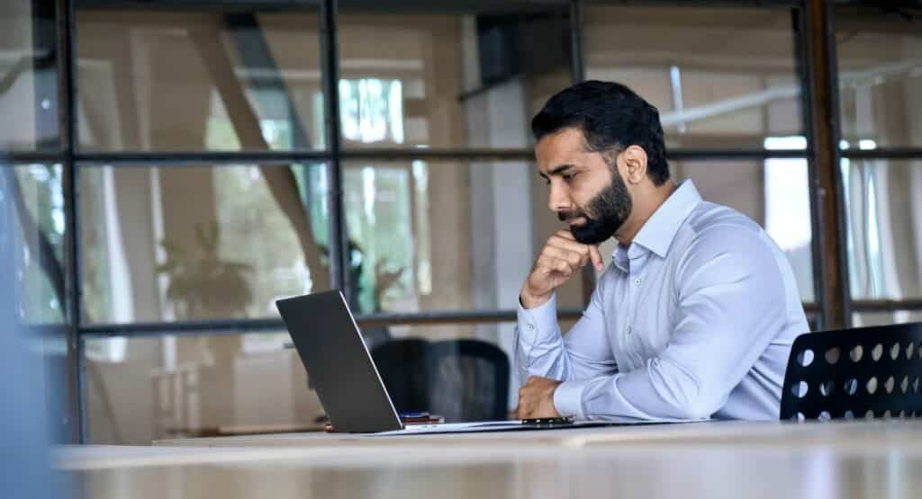 Focused man with a beard working intently on a laptop in a modern office setting.