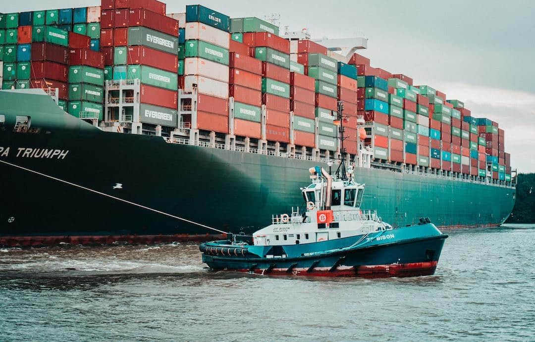 Tugboat "Bison" in blue and red colors guiding the massive "Evergreen" container ship in a busy port.