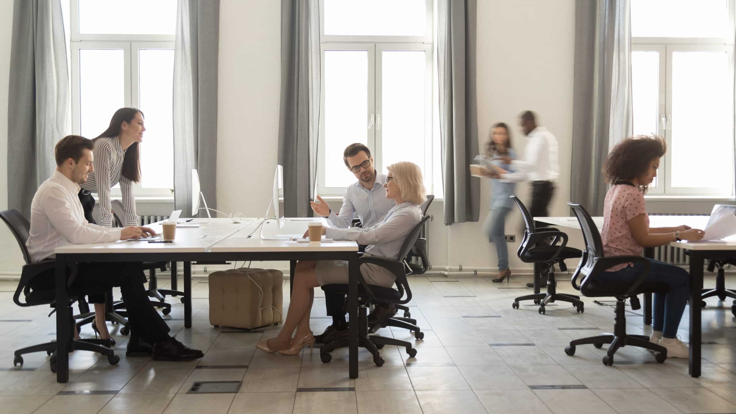 A diverse group of business professionals in a meeting room with one person pointing at a document while another stands and looks on. Blurred figures in the background suggest a dynamic office setting.