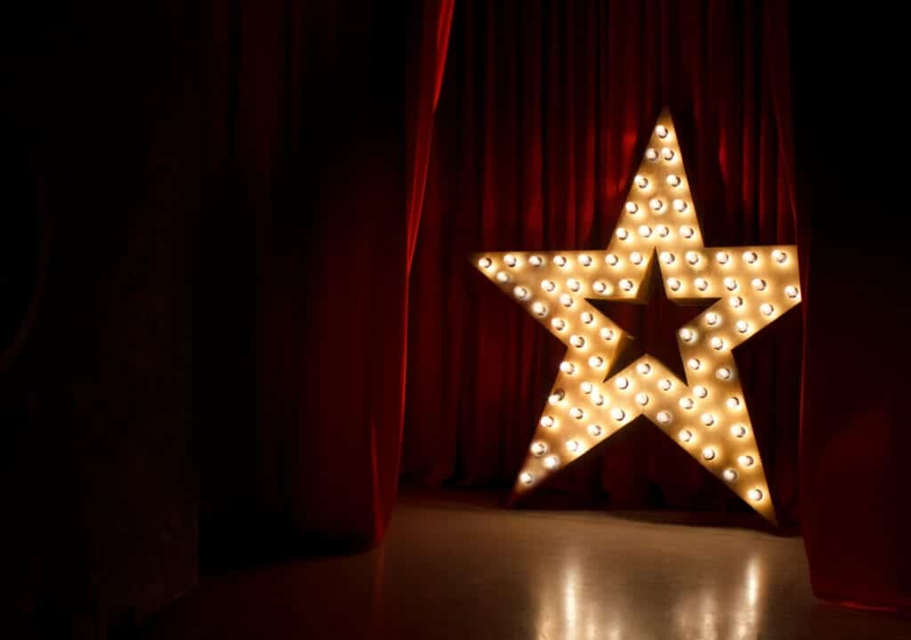 Brightly lit star with light bulbs in front of a red velvet curtain backdrop.