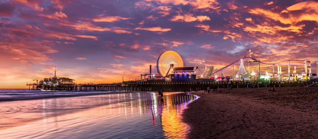 A vibrant sunset paints the sky in shades of pink and orange above the Santa Monica Pier, with the Ferris wheel and roller coaster illuminated against the evening light