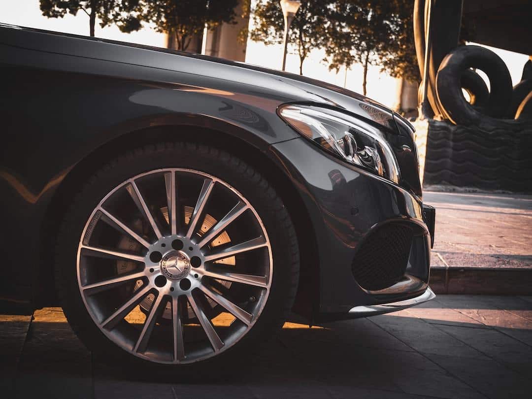 Close-up of a black luxury car's front wheel and grille, parked beside trees at sunset.