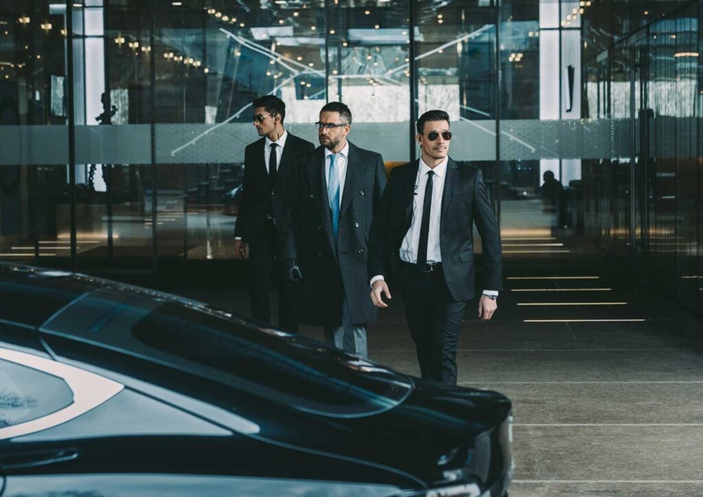 Understanding The Core Roles And Responsibilities Of A Close Protection Agent 4 Three close protection agents in suits, walking confidently outside a modern office building, showcasing professionalism and security services.