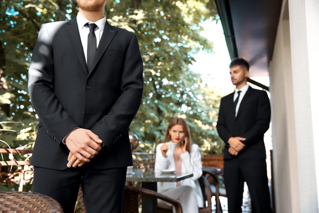 Understanding The Core Roles And Responsibilities Of A Close Protection Agent 5 A close protection agent standing guard near a young businesswoman seated at an outdoor café, providing discreet security services.