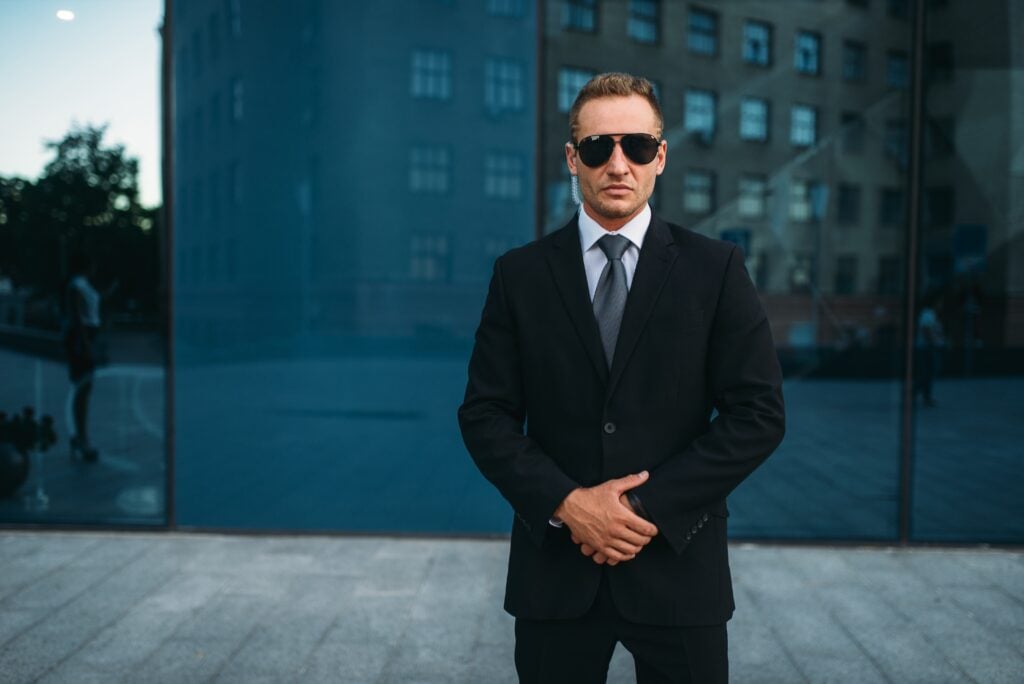 The Ultimate Guide To Close Protection Services: Safeguarding High-Profile Lives With Precision And Expertise 9 Professional bodyguard in a suit with sunglasses and earpiece standing confidently outside a modern building, representing close protection services.