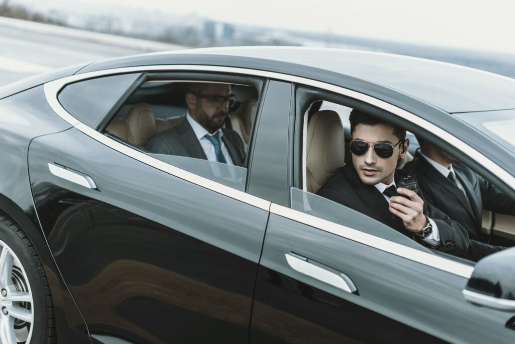 Businessman sitting in the back seat of a luxury black car with personal security details providing discreet protection.