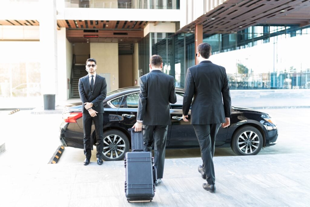 Close protection for CEOs showing bodyguards escorting a VIP with a suitcase, leaving a modern corporate building in a black car.