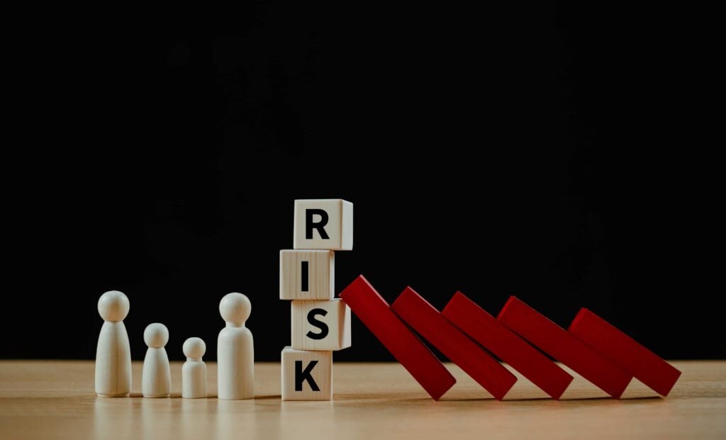 Risk management concept represented by falling red dominoes, wooden figures, and letter blocks spelling "RISK," symbolizing crisis prevention and protection.
