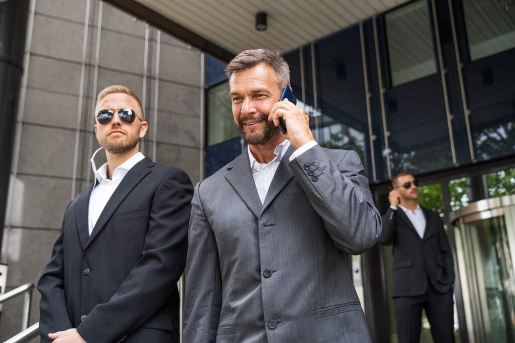 A businessman on the phone being protected by two professional security guards in suits standing outside a modern office building.