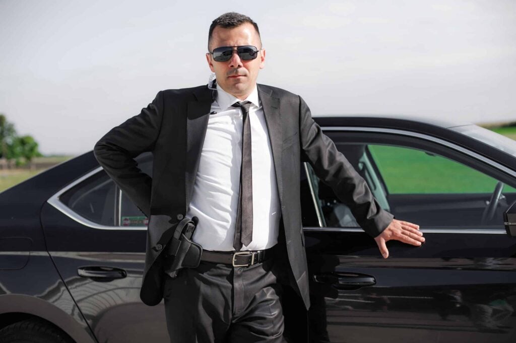 A professional bodyguard in a suit and sunglasses standing beside a luxury car, equipped for Murfreesboro Bodyguard Services.