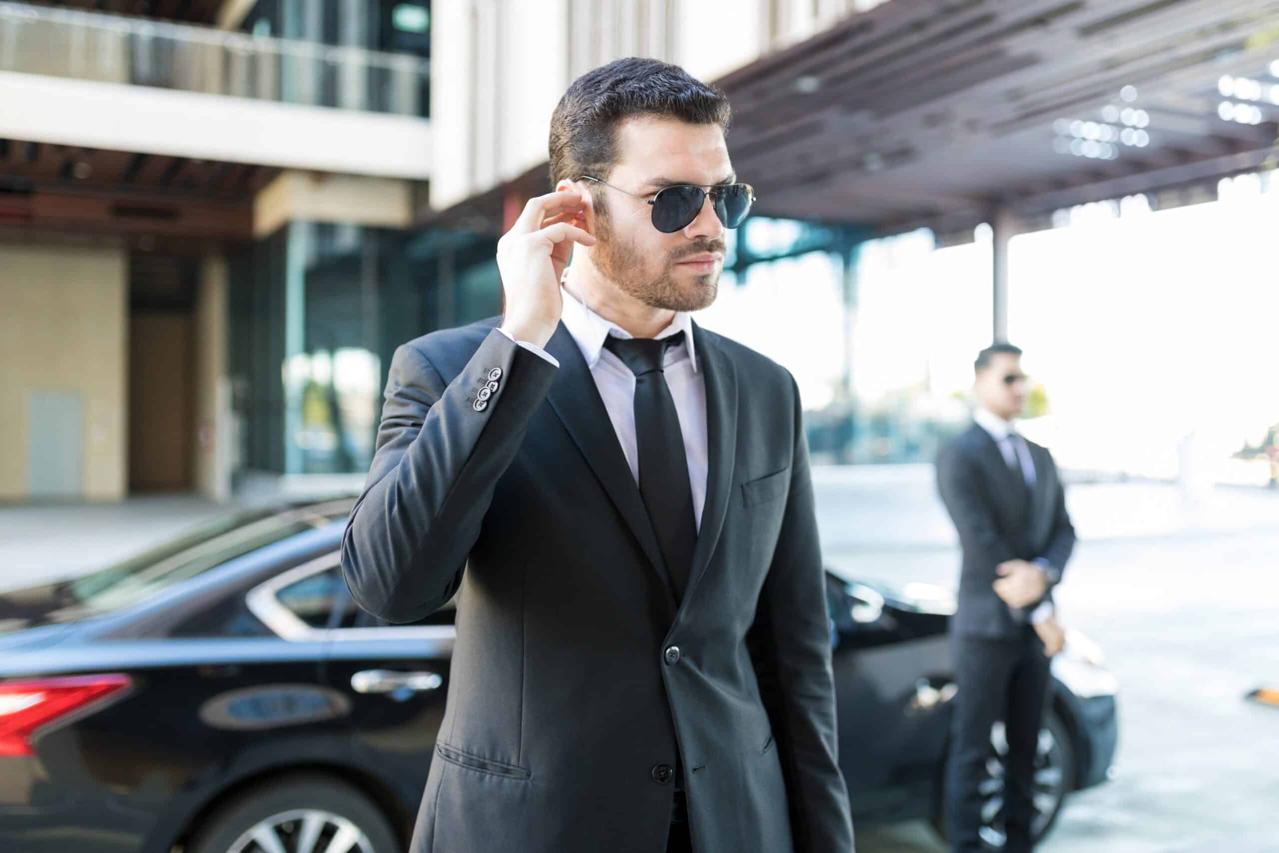 Male security agent with earpiece standing alert by black sedan, illustrating the Seven Principles of Close Protection