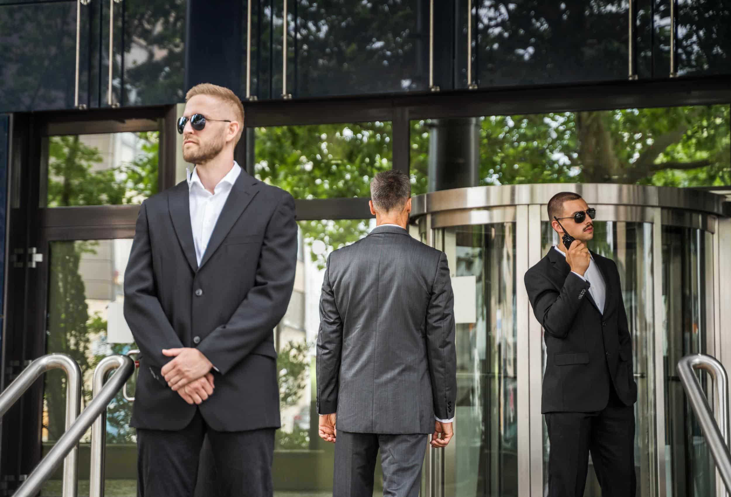 Three suited security professionals standing alert outside a modern glass building during Close Protection Operational Planning.