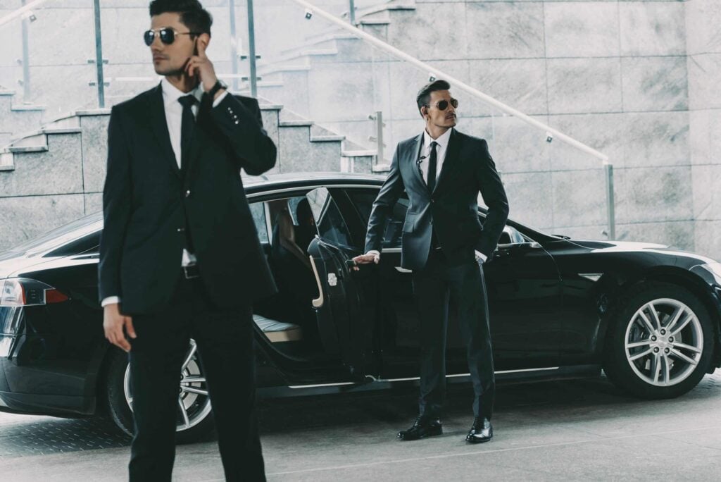 Close Protection Journey Management with two security agents escorting a client from a black vehicle outside a modern office building.