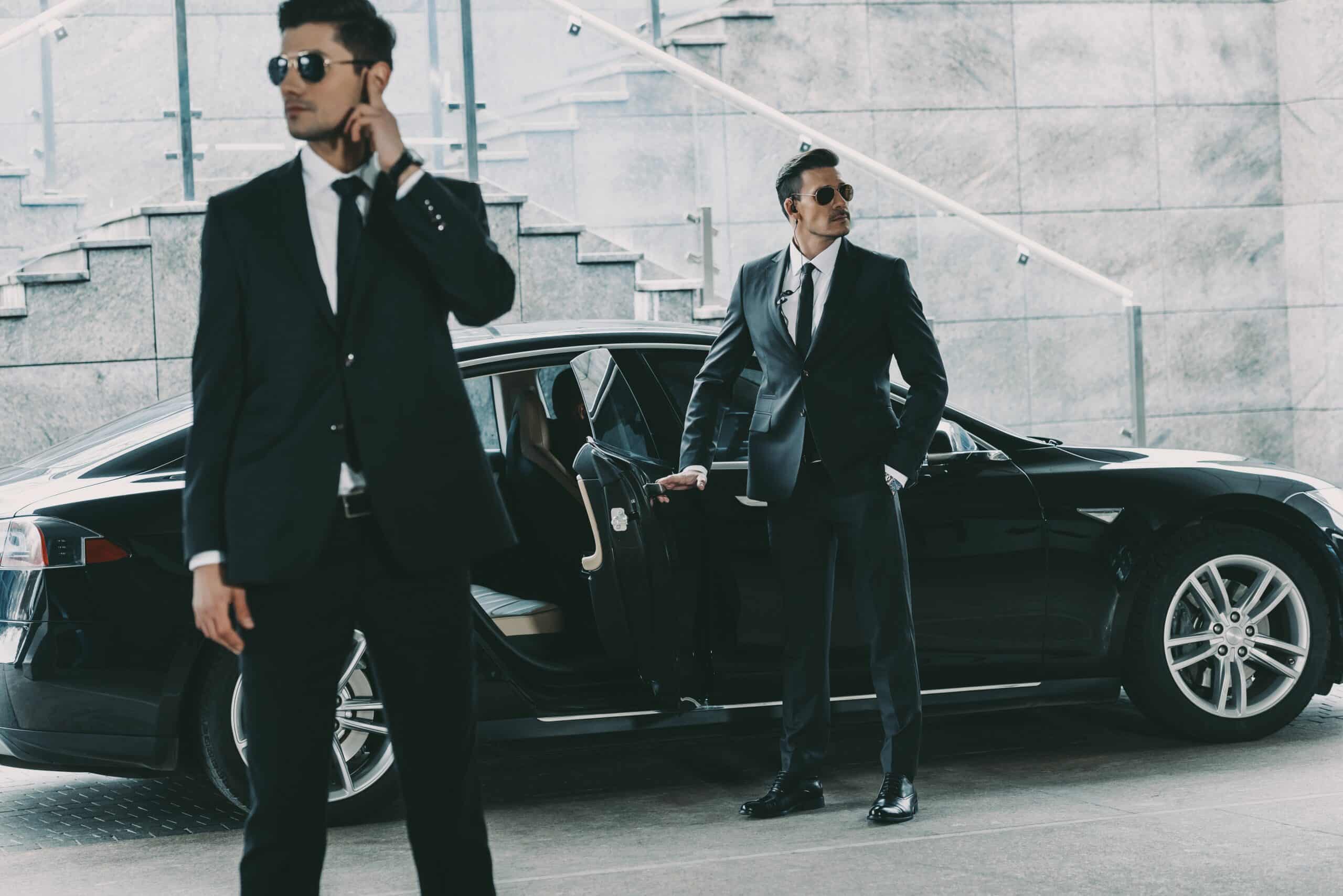 Close Protection Journey Management with two security agents escorting a client from a black vehicle outside a modern office building.
