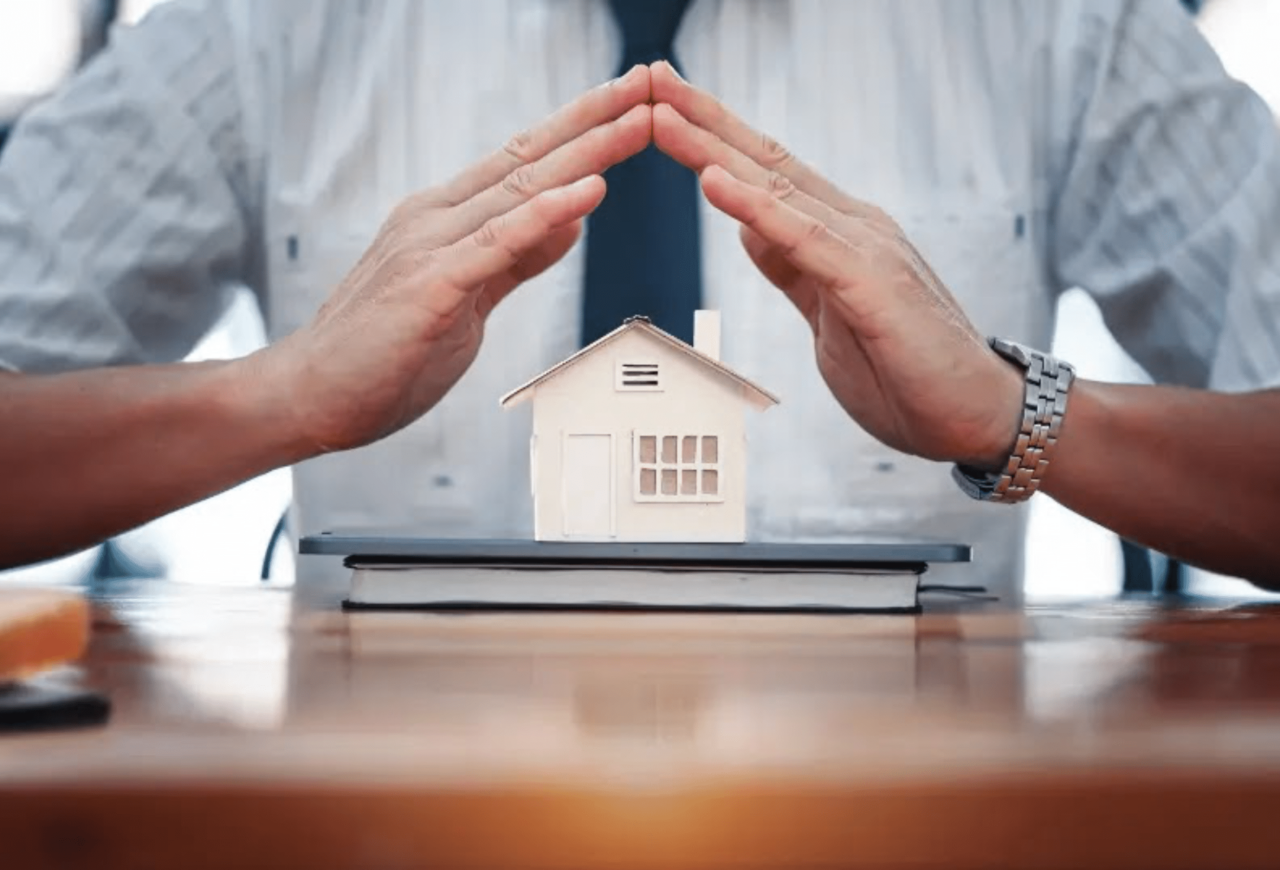 Businessman’s hands forming a protective roof gesture over a miniature house model on a table, symbolizing protection against threats to private estates.