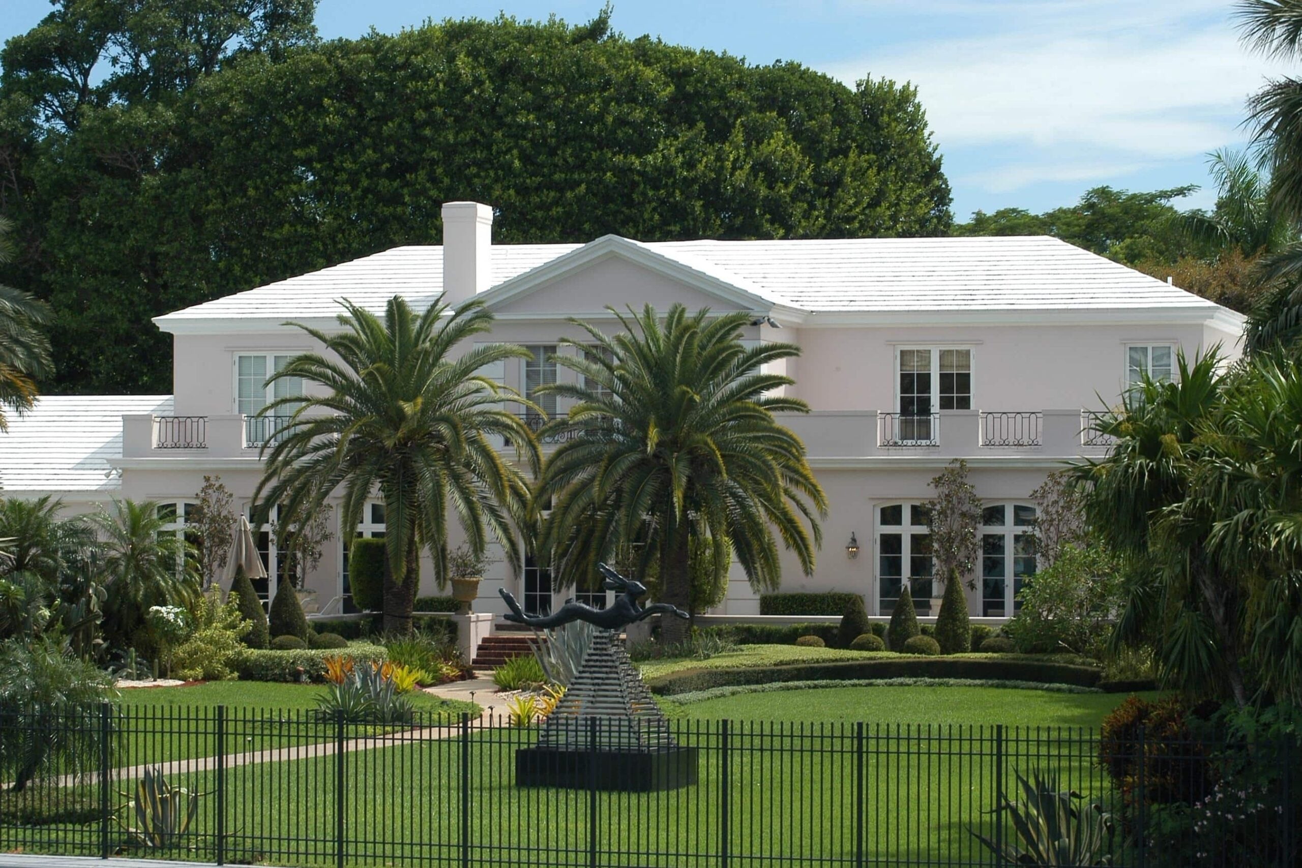 Elegant white luxury estate with palm trees and wrought iron fence requiring property protection while traveling security services