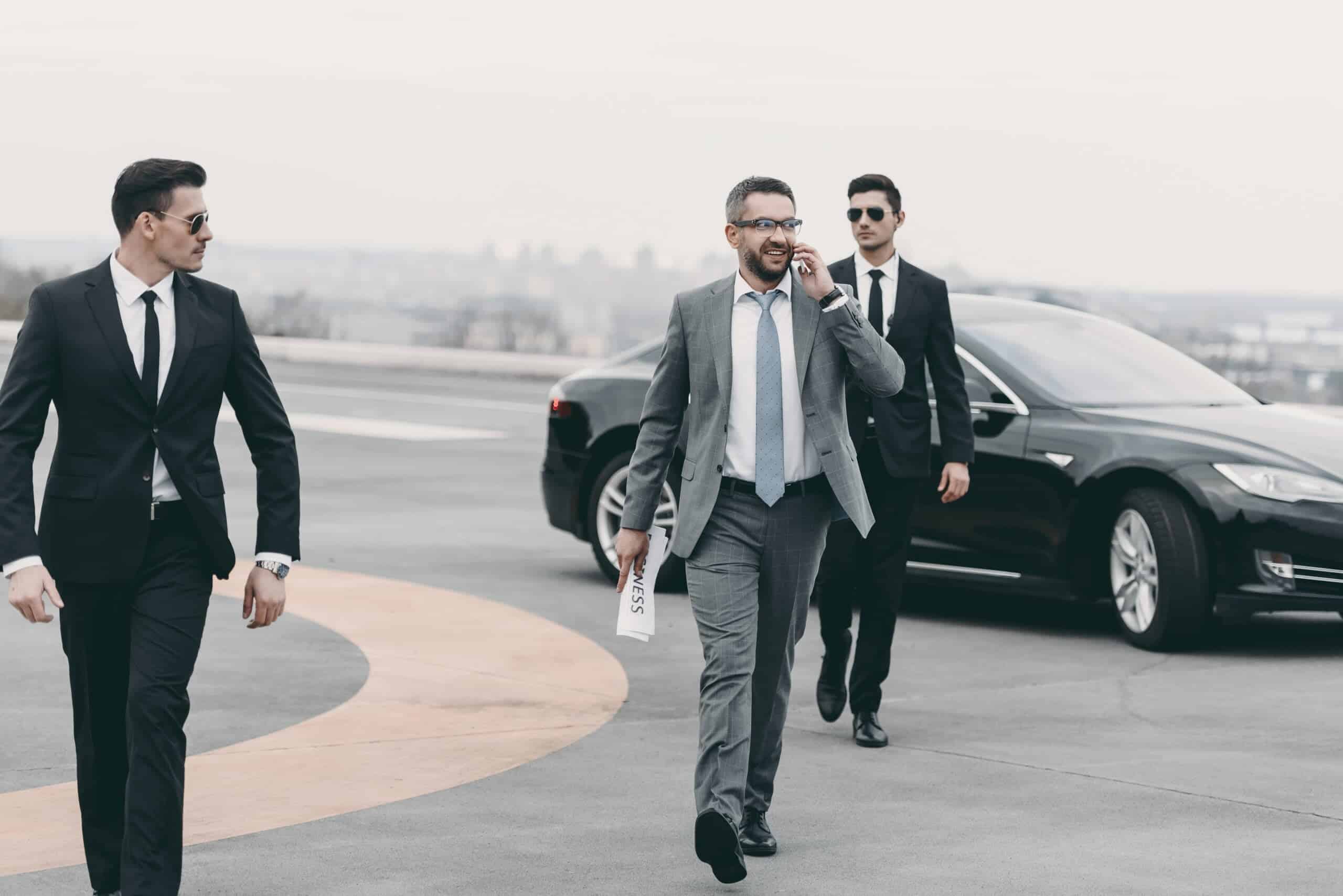 A suited executive talks on his phone while walking across a rooftop helipad flanked by two close protection agents in dark suits and sunglasses beside a black luxury sedan, illustrating the airport arrival and departure security risk management required for high-profile principals