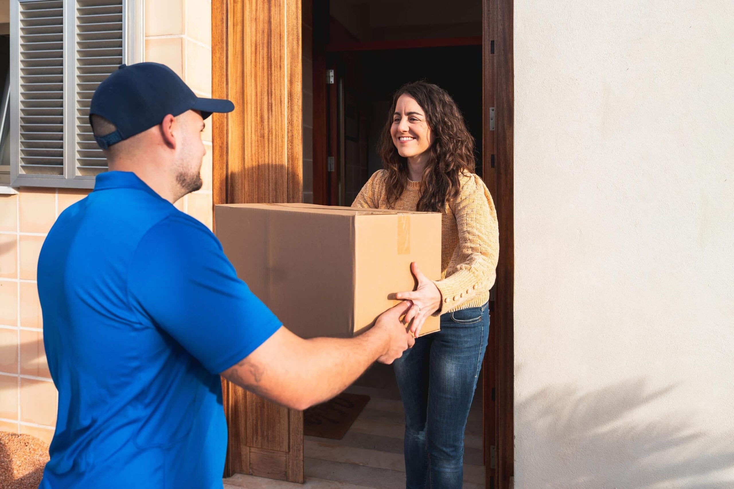 A smiling woman opens her front door to accept a large cardboard box from a delivery person in a blue uniform, highlighting the need for vendor and delivery screening security at residential properties