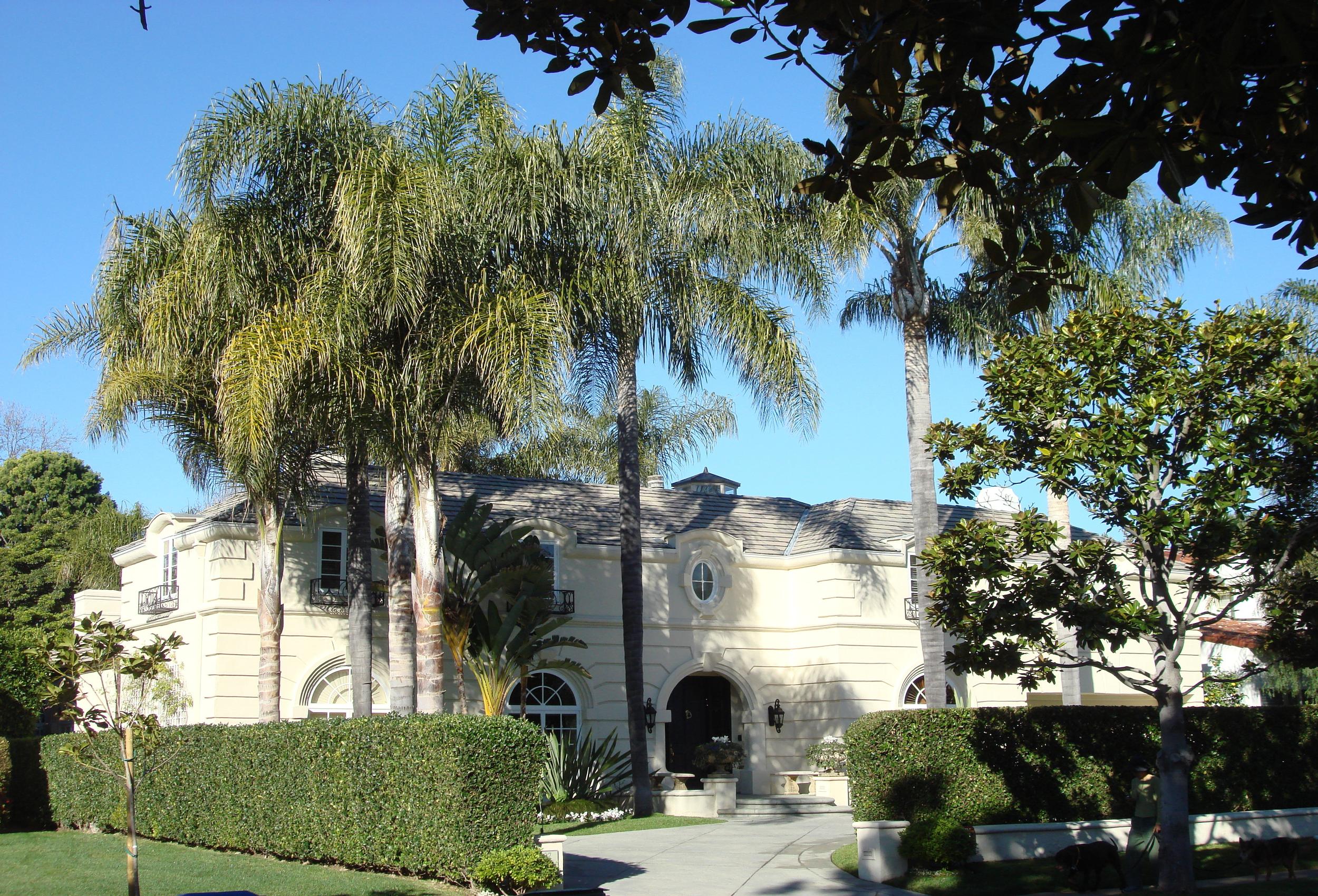 A street-level view of a grand cream-colored luxury estate with a French-inspired facade, arched entry, tall queen palms, a neatly clipped hedge perimeter, and mature magnolia trees along the front boundary, illustrating the layered vegetation challenges of estate security for outdoor living spaces at high-value residential properties
