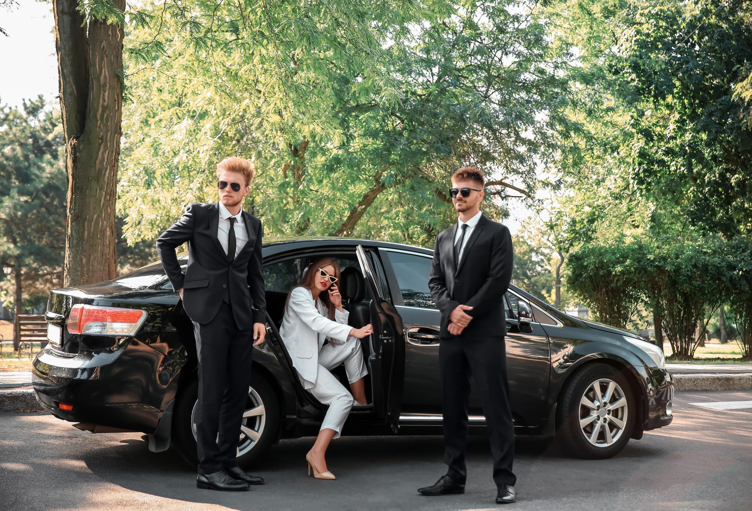 Two professional bodyguards in black suits stand guard beside a black luxury sedan as a female VIP in a white suit exits the vehicle in a tree-lined outdoor setting, demonstrating how open-environment vehicle exits represent significant executive security vulnerabilities