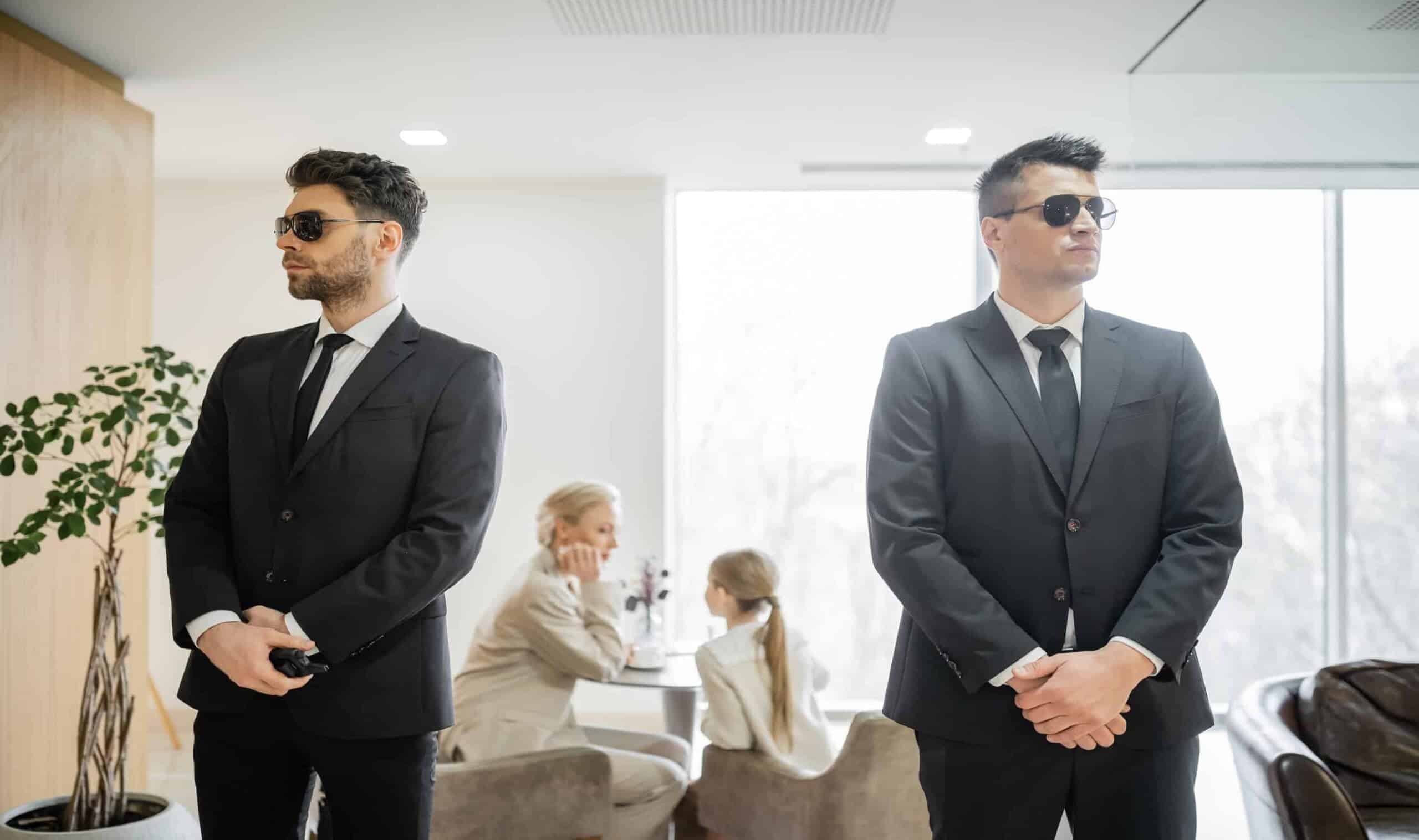 Two suited security agents wearing sunglasses stand at attention in a bright private lounge while a woman and child sit at a table behind them, demonstrating the protective environment created through generational wealth security planning