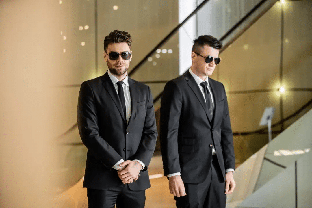 Two close protection agents in black suits and aviator sunglasses stand in alert formation at the base of a curved staircase inside a warm, upscale interior space, representing the intelligence led executive protection deployment used to secure high-value venue environments